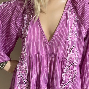 Puede incluir: Vestido a rayas moradas y blancas con cuello en V y mangas largas. El vestido presenta bordados florales blancos en la parte delantera y en las mangas. El vestido tiene una falda escalonada y un corte holgado.