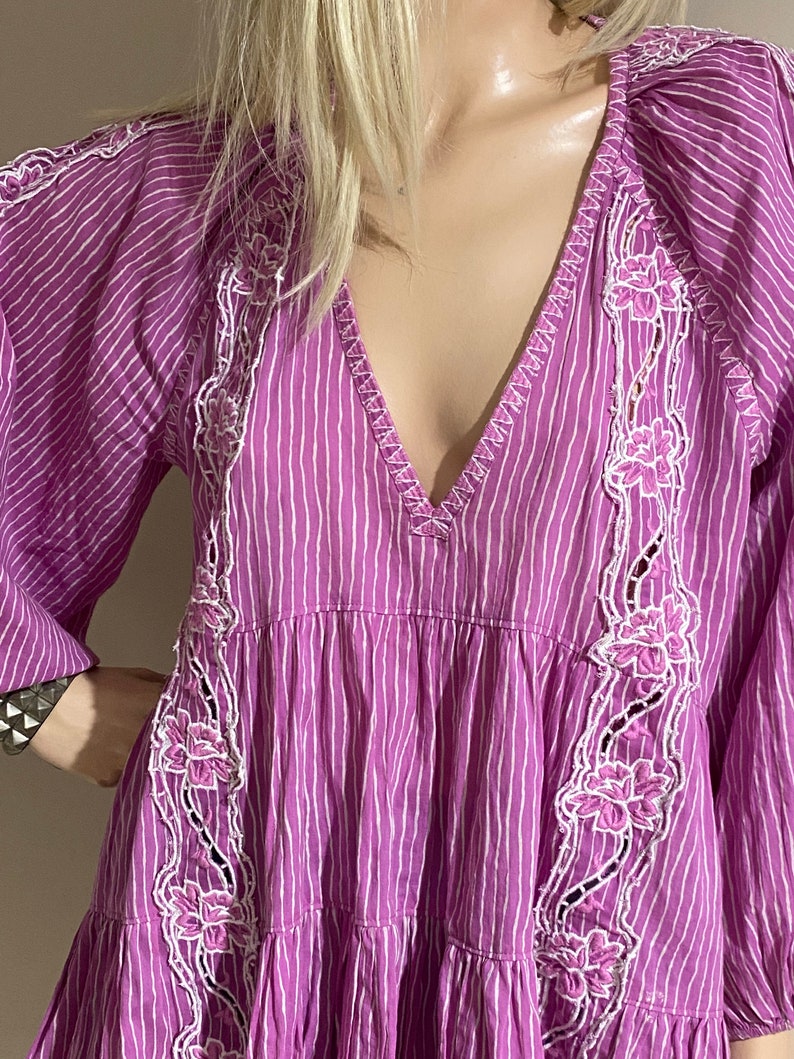 Puede incluir: Vestido a rayas moradas y blancas con cuello en V y mangas largas. El vestido presenta bordados florales blancos en la parte delantera y en las mangas. El vestido tiene una falda escalonada y un corte holgado.