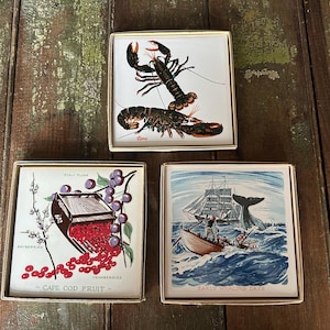 May include: Three square coasters with gold-colored metal frames. One coaster features a lobster illustration, another shows cranberries with the text "Cape Cod Fruit", and the third depicts a whaling scene with the text "Early Whaling Days".