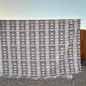 May include: A large white textile with a repeating geometric pattern of diamonds and hexagons in shades of brown, black, and pink. The textile is hanging on a clothesline outdoors, with a wooden fence in the background.