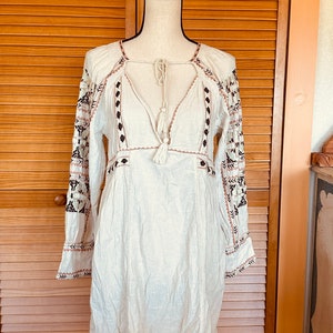 May include: A white, long-sleeved dress with intricate embroidery. The dress features a V-neckline with a tassel tie, and the sleeves are adorned with black and brown floral patterns. The dress is displayed on a mannequin.