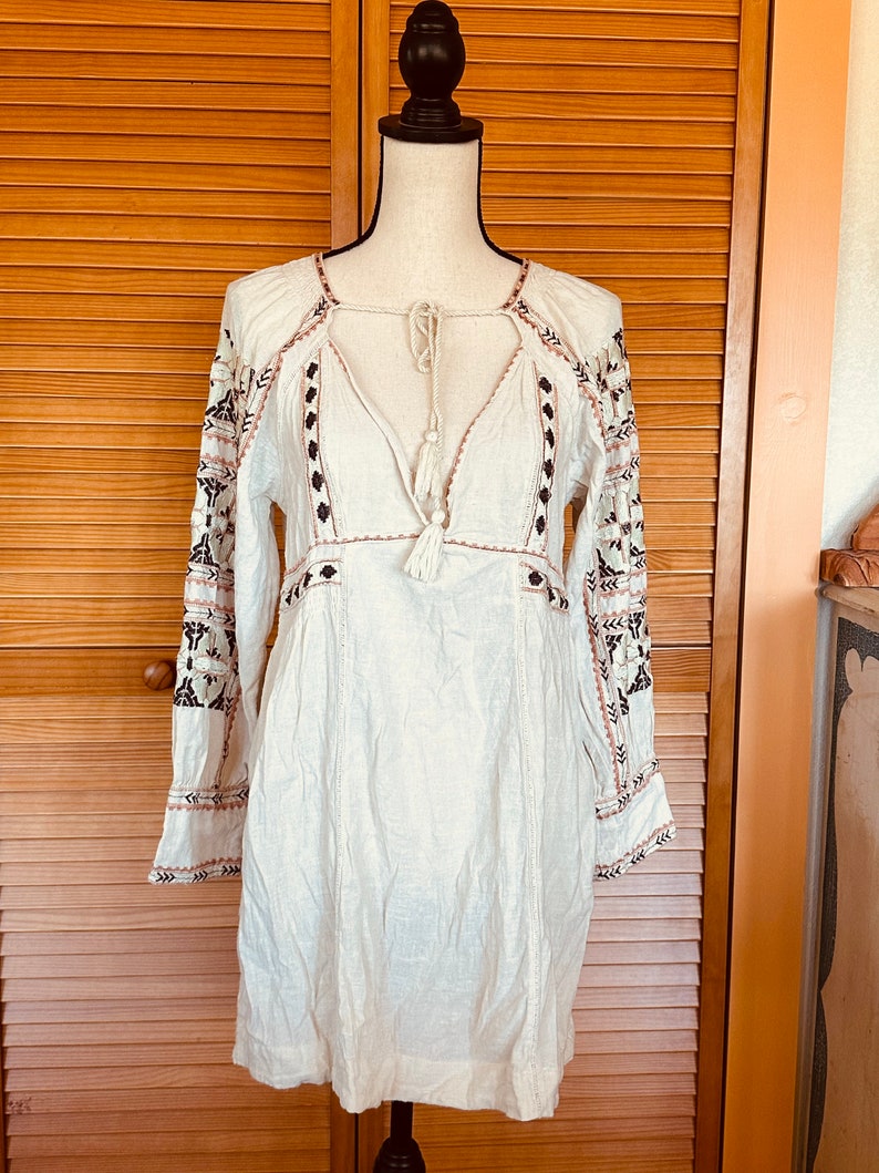 May include: A white, long-sleeved dress with intricate embroidery. The dress features a V-neckline with a tassel tie, and the sleeves are adorned with black and brown floral patterns. The dress is displayed on a mannequin.