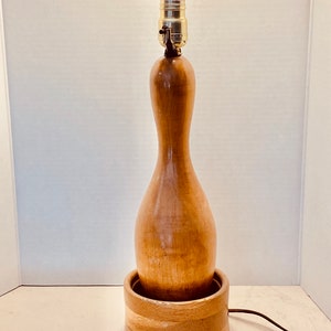 May include: A table lamp featuring a light brown wooden bowling pin as the central body, set into a circular wooden base. The lamp includes a metallic socket at the top, a white light bulb, and a thin brown electrical cord extending from the side.