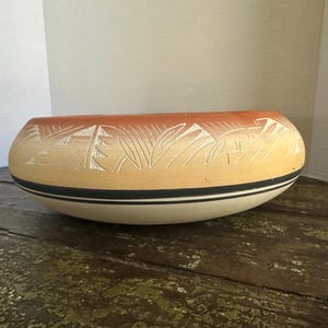 May include: A low-profile ceramic bowl featuring a gradient color scheme transitioning from terracotta orange at the rim to a pale cream base. The exterior is decorated with carved geometric patterns and two thin black horizontal lines near the bottom.