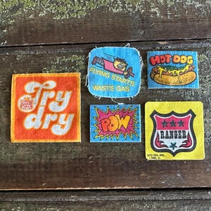 May include: A collection of five vintage fabric patches with various designs. One patch features the text "Try dry" in orange and white. Other patches include a hot dog, a plane, the word "Pow", and a ranger badge.