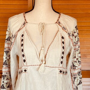 May include: Cream-colored long-sleeve tunic with embroidered details. The tunic features a square neckline, a V-neck with a tassel tie, and intricate embroidery in brown, black, and pink. The sleeves also have embroidered patterns.