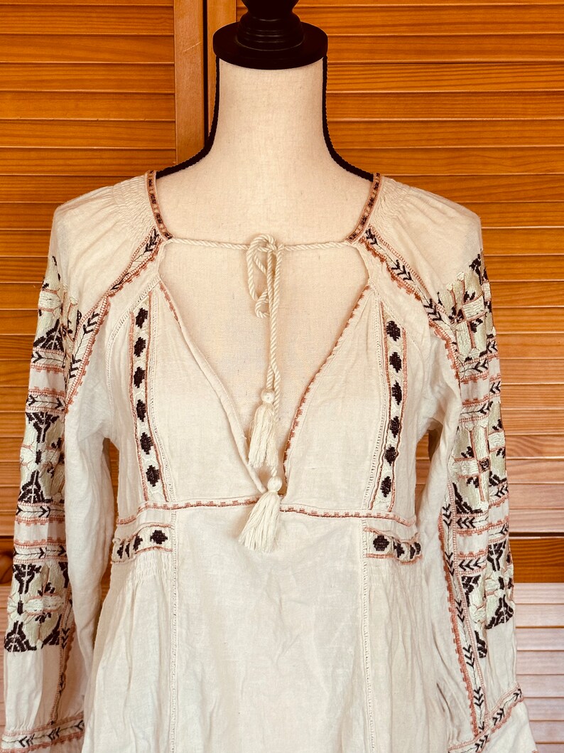 May include: Cream-colored long-sleeve tunic with embroidered details. The tunic features a square neckline, a V-neck with a tassel tie, and intricate embroidery in brown, black, and pink. The sleeves also have embroidered patterns.