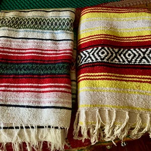 May include: Two folded woven blankets with fringe edges. The left blanket features horizontal stripes in white, red, green, and black with a diamond pattern. The right blanket displays horizontal stripes in yellow, white, and red with a black diamond pattern.