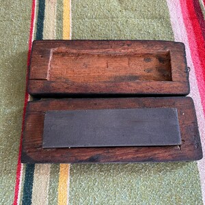 Op de afbeelding: Een open, antieke houten kist met een donkere, gebeitste afwerking. Binnenin is een rechthoekige, donkergrijze slijpsteen zichtbaar. De kist ligt op een gestreepte, veelkleurige stof.