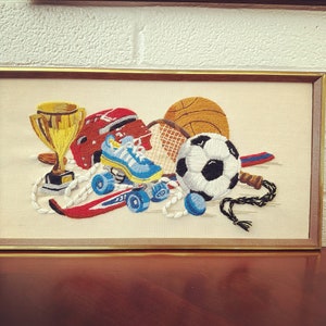 May include: A rectangular framed embroidery featuring a collection of sports equipment including a gold trophy, a red helmet, a blue sneaker, a basketball, a soccer ball, a tennis racket, a red ski, and a jump rope on a cream-colored fabric background.