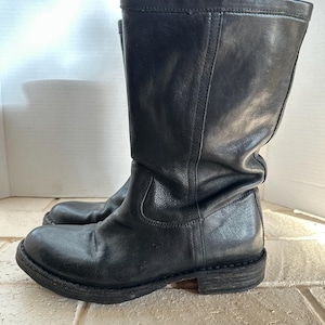 May include: A pair of black leather boots. The boots have a mid-calf height, a rounded toe, and a low heel. The leather appears worn, with visible creases and a matte finish. The soles are dark and show signs of wear.