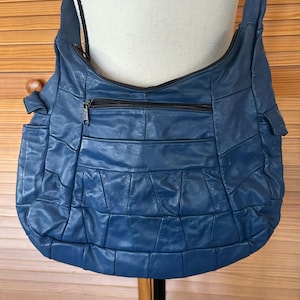 May include: A blue leather shoulder bag with a quilted design. The bag has a long strap, a zippered main compartment, and an exterior pocket. The bag is displayed on a mannequin.