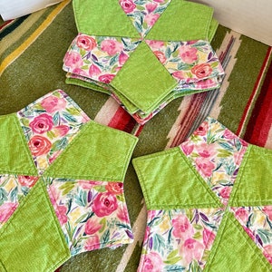 May include: A stack of star-shaped coasters. Each coaster features a vibrant floral pattern with pink roses and green leaves, contrasted by solid green fabric. The coasters are arranged on a striped rug.