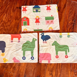 May include: Three white pillowcases with colorful illustrations. One pillowcase features houses, figures, and a barn. The other two pillowcases have animal figures with wheels. The animals include a camel, horse, and a bird.