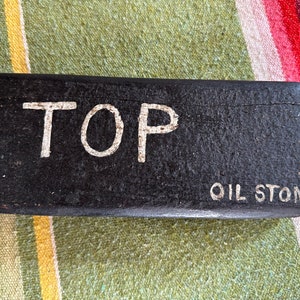 Op de afbeelding: Een rechthoekige zwarte olieslijpsteen met het woord "TOP" in witte hoofdletters. Onder het woord "TOP" staat de tekst "OIL STONE" in witte hoofdletters. De steen ligt op een gestreept oppervlak.