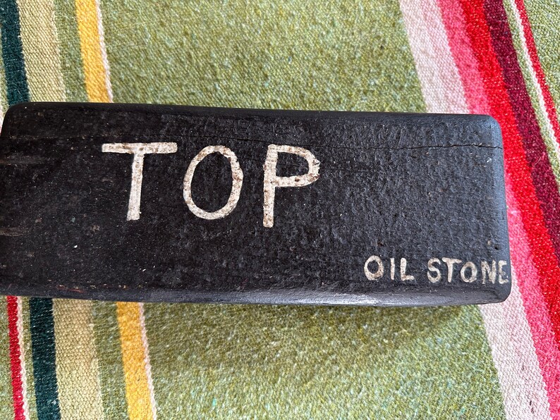 Op de afbeelding: Een rechthoekige zwarte olieslijpsteen met het woord "TOP" in witte hoofdletters. Onder het woord "TOP" staat de tekst "OIL STONE" in witte hoofdletters. De steen ligt op een gestreept oppervlak.