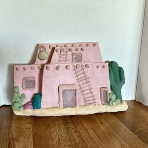 May include: A pink Southwestern-style decorative wall hanging. The piece depicts a pueblo-style building with a ladder, windows, and a cactus. The building is painted in a light pink color with a textured finish. The base is a light tan color.