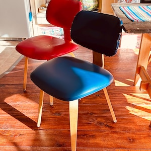 May include: Three mid-century modern chairs with wooden legs and colorful seats. One chair has a red seat and back, another has a navy blue seat and back, and the third has a blue seat and a black back. The chairs are on a wooden floor.