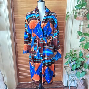 May include: A patterned robe in shades of brown, blue, and orange, displayed on a mannequin. The robe features a shawl collar, long sleeves, and a matching belt. The design includes geometric shapes and stripes.