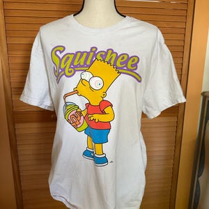 May include: White t-shirt featuring Bart Simpson drinking a "Squishee" drink. The graphic shows Bart in a red shirt, blue shorts, and blue shoes. The word "Squishee" is in purple and yellow lettering.