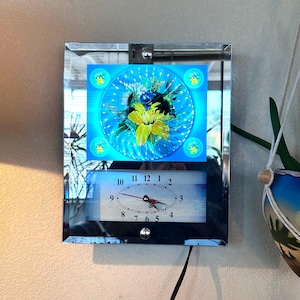 May include: A rectangular wall clock with a mirrored frame featuring a central circular graphic of a yellow lily and a hummingbird against a bright blue background. Below the graphic is an oval clock face with black numbers and red hands.
