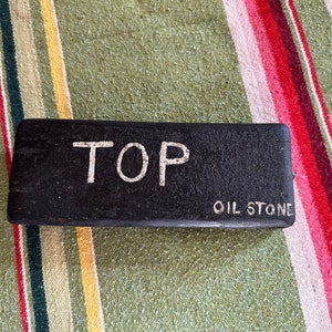 Op de afbeelding: Een rechthoekige zwarte olie-steen met de woorden "TOP" en "OIL STONE" in witte letters. De steen ligt op een gestreepte achtergrond van groen, geel, rood en wit.
