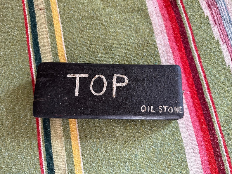 Op de afbeelding: Een rechthoekige zwarte olie-steen met de woorden "TOP" en "OIL STONE" in witte letters. De steen ligt op een gestreepte achtergrond van groen, geel, rood en wit.
