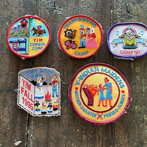 May include: Five embroidered fabric patches featuring various designs. One circular patch reads YIM COWGIRL CAMP, one circular patch reads THANKSGIVING CAMP 96, one rectangular patch reads SOCK HOP CAMP 97, one shield-shaped patch reads STORY BOOK CAMP YIM FALL 1995, and one circular patch reads Y-INDIAN MAIDENS MOTHER DAUGHTER FRIENDS ALWAYS.