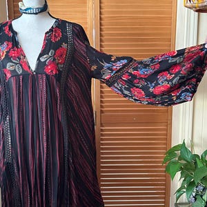 May include: Black dress with red and black vertical stripes and floral print sleeves. The dress has a V-neck and lace detailing. The sleeves feature a vibrant floral pattern with red, blue, and yellow flowers. The dress is displayed on a mannequin.