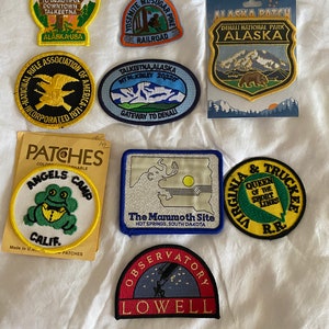 May include: A collection of embroidered patches featuring various designs and text. The patches include images of a frog, a bear, mountains, and text such as "Alaska," "Yosemite," and "Angels Camp, Calif." Some patches are on cards.