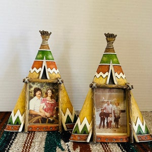 May include: Two decorative teepee-shaped picture frames. The frames are painted with a Native American design in yellow, green, and orange. Each frame holds a photograph. The frames are resting on a patterned blanket.