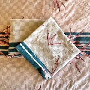 May include: Bedding set featuring a tan and white checkered pattern with a teal and white striped border. The set includes a folded pillowcase and a sheet, both adorned with stylized maroon leaf designs.