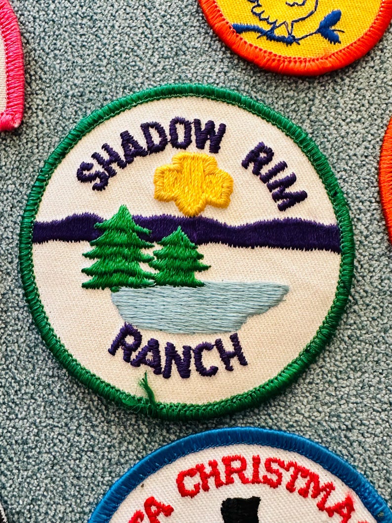 Op de afbeelding: Geborduurde patch met de woorden "SHADOW RIM RANCH" in marineblauw. De patch toont een gele zon, twee groene dennenbomen en een blauw meer tegen een witte achtergrond, omlijst door een groene rand.