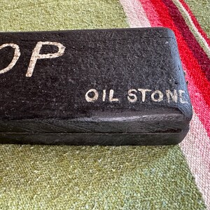 Op de afbeelding: Een zwarte rechthoekige olie-steen met de woorden "TOP" en "OIL STONE" in witte letters. De steen heeft een getextureerd oppervlak. De achtergrond is een kleurrijke gestreepte stof.