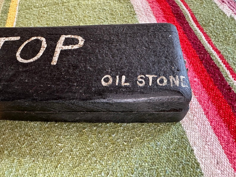 Op de afbeelding: Een zwarte rechthoekige olie-steen met de woorden "TOP" en "OIL STONE" in witte letters. De steen heeft een getextureerd oppervlak. De achtergrond is een kleurrijke gestreepte stof.