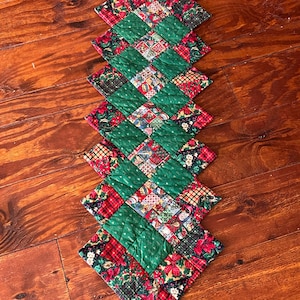 May include: A quilted Christmas table runner with a diamond pattern. The runner features alternating green squares and squares with a patchwork of red, green, and white holiday-themed fabrics. The runner is approximately 40 inches long.