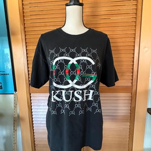 May include: Black t-shirt with a white, red, and green graphic design. The design includes the letters "OG" and the word "KUSH" in white block letters. The shirt is displayed on a mannequin.