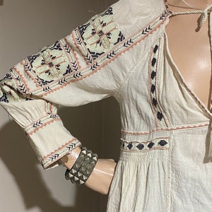 May include: Cream-colored long-sleeve dress with intricate embroidered details in shades of blue, peach, and green. The dress features a V-neckline and a drawstring closure. A silver studded bracelet is visible on the mannequin's arm.