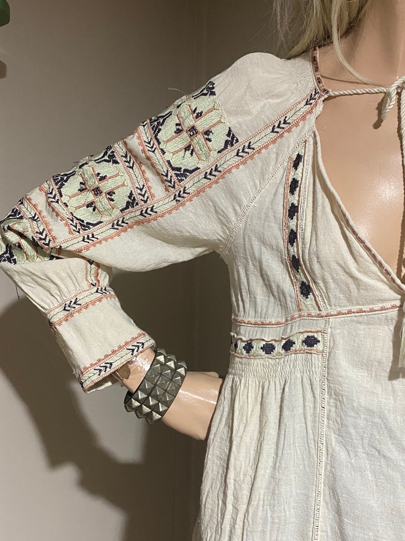 May include: Cream-colored long-sleeve dress with intricate embroidered details in shades of blue, peach, and green. The dress features a V-neckline and a drawstring closure. A silver studded bracelet is visible on the mannequin's arm.