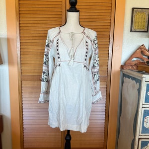 May include: A white, long-sleeved dress with intricate embroidery on the sleeves and neckline. The dress features a V-neck with a tassel tie and decorative stitching. The dress is displayed on a black mannequin against a wooden door background.