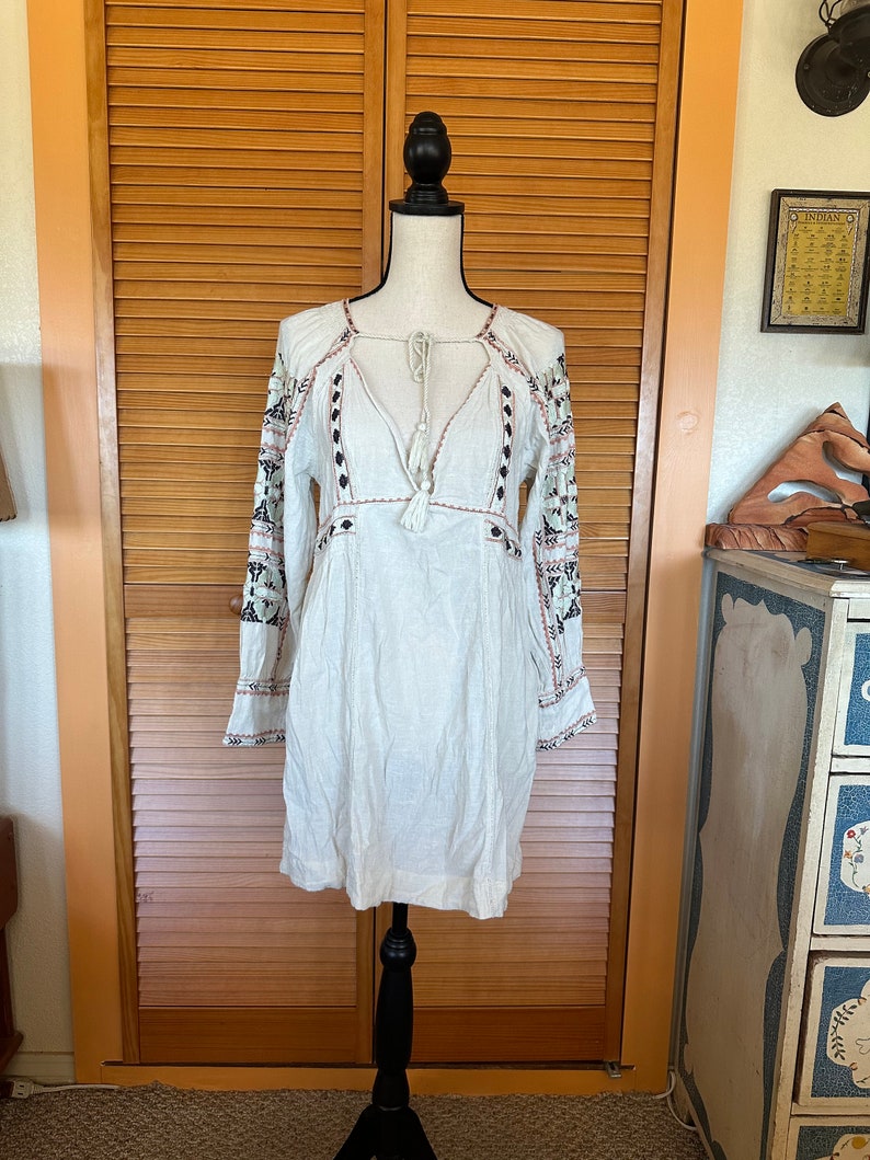 May include: A white, long-sleeved dress with intricate embroidery on the sleeves and neckline. The dress features a V-neck with a tassel tie and decorative stitching. The dress is displayed on a black mannequin against a wooden door background.
