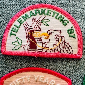 Op de afbeelding: Geborduurde patch met de tekst "TELEMARKETING '87" in groen, gebogen aan de bovenkant. De patch heeft een bruine en gele illustratie van een telefoon en een vogel. De patch heeft een roze rand.