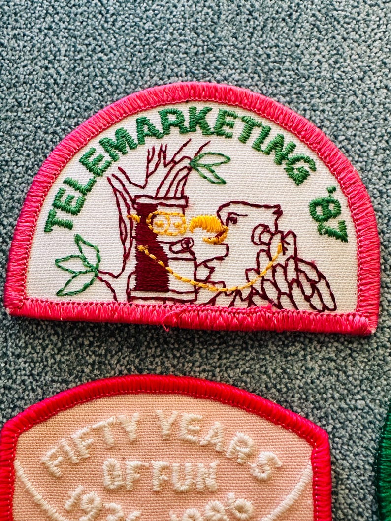 Op de afbeelding: Geborduurde patch met de tekst "TELEMARKETING '87" in groen, gebogen aan de bovenkant. De patch heeft een bruine en gele illustratie van een telefoon en een vogel. De patch heeft een roze rand.