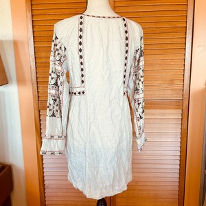 May include: Cream-colored tunic dress with long sleeves and intricate embroidery. The dress features detailed patterns in black, brown, and pink along the sleeves, neckline, and bodice. The dress is displayed on a black mannequin.