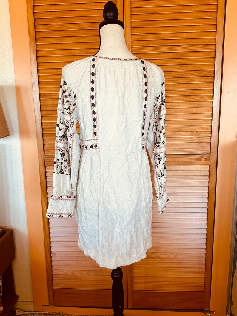 May include: Cream-colored tunic dress with long sleeves and intricate embroidery. The dress features detailed patterns in black, brown, and pink along the sleeves, neckline, and bodice. The dress is displayed on a black mannequin.