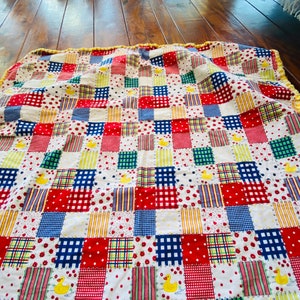 May include: A patchwork quilt with a white background and squares of red, blue, yellow, and green fabrics. The quilt features patterns like polka dots, stripes, and gingham, with yellow duck appliqués. The quilt has a yellow fringe.