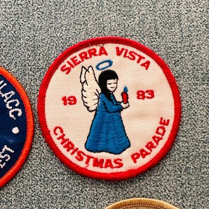 Op de afbeelding: Een ronde patch met een rode rand, met de tekst "SIERRA VISTA CHRISTMAS PARADE" en de jaartallen "1983" en "19". Het toont een engel in een blauwe jurk die een kaars vasthoudt, met zwart haar en een halo.