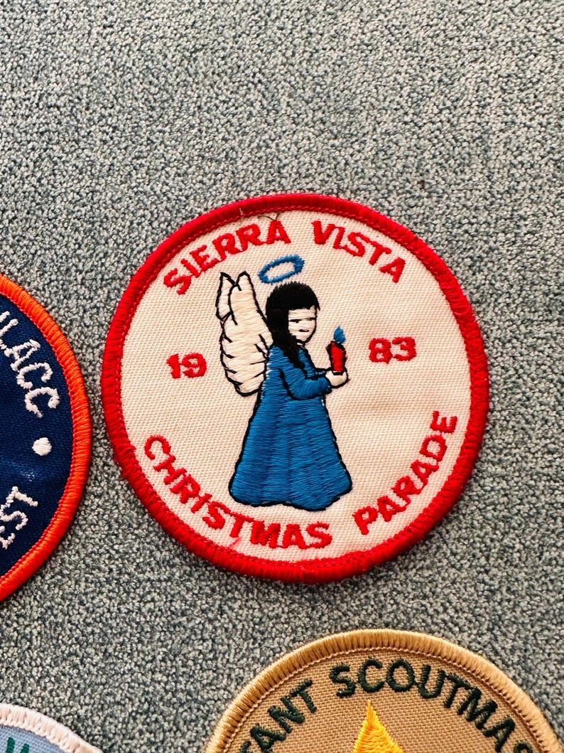 Op de afbeelding: Een ronde patch met een rode rand, met de tekst "SIERRA VISTA CHRISTMAS PARADE" en de jaartallen "1983" en "19". Het toont een engel in een blauwe jurk die een kaars vasthoudt, met zwart haar en een halo.