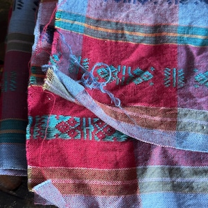 May include: Close-up of a colorful woven textile with a plaid pattern. The fabric features a combination of red, blue, and teal hues, with intricate geometric designs. The textile appears to be handmade, with visible threads and a slightly frayed edge.