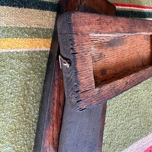 Op de afbeelding: Een verweerd, donkerbruin houten gereedschap met een rechthoekige basis en een verhoogd, open bovendeel. Het hout vertoont tekenen van ouderdom en slijtage, met zichtbare nerf en een metalen sluiting. Het gereedschap rust op een groen, rood en geel gestreept textiel.